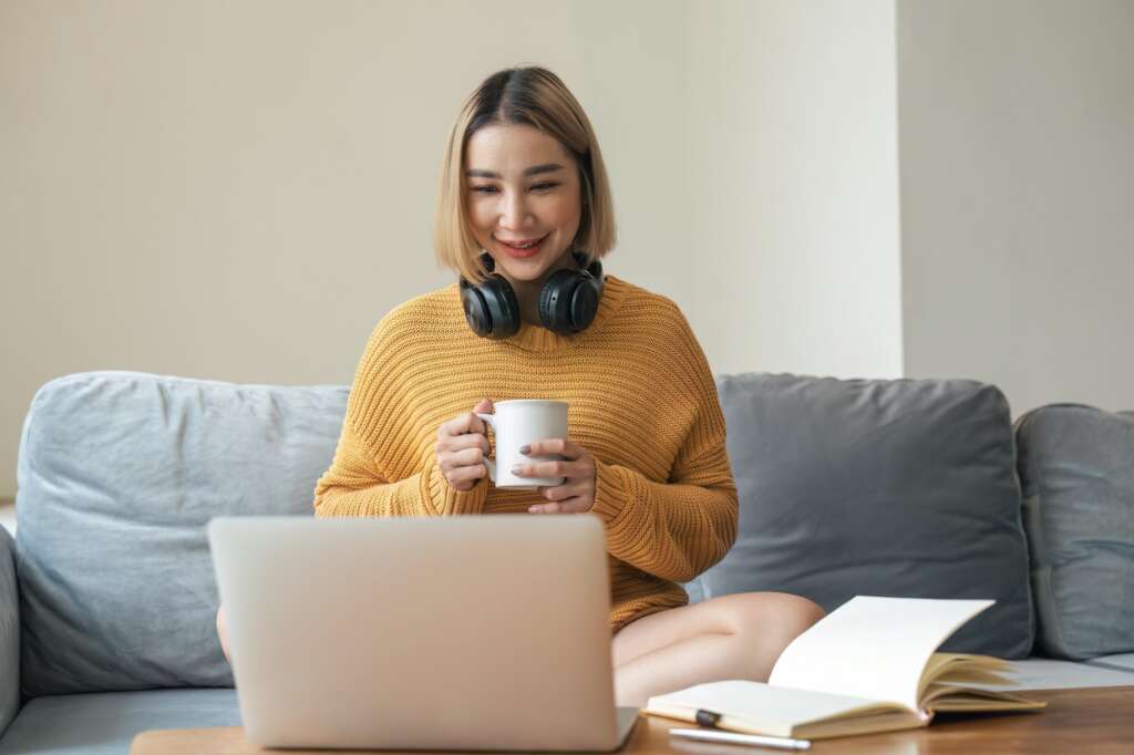 Mulher asiática, com cabelos loiros, blusa amarela e uma xícara branca na mão, em frente ao um laptop. Participando de uma sessão de terapia online para brasileiras no exterior  